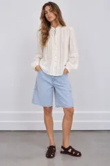 Blusa de tejido texturizado color blanco, con cuello alto con volados y cordón para atar, cierre frontal de botones y mangas largas abullonadas con detalles de encaje y tablas verticales.