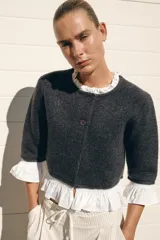 Chaqueta gris de cuello redondo y manga tres cuartos, con detalle de volantes de popelín blanco en el cuello, puños y bajo. Cierre frontal con botones.