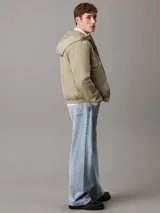 Campera Calvin Klein color verde oliva, con capucha, cierre frontal con cremallera, puños y bajo elásticos, bolsillos con botón de presión en la cintura y logo de la marca en el pecho y la manga. Confeccionada en nailon 100% reciclado y forro de poliéster.