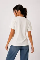 Remera de algodón color blanco off-white, de manga corta, cuello redondo a la base y corte holgado. Presenta un estampado frontal con diseño de corazón en tonos marrones y azules, con detalles de strass y texto.