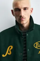 Cazadora estilo varsity con cuerpo de mezcla de lana en color verde oscuro y mangas de efecto piel a tono. Presenta cuello solapa, cierre frontal con botones a presión, bolsillos laterales de vivo y puños y bajo acanalados con rayas amarillas. Incluye bordados y parches decorativos en el frente con la inscripción Boston Archive.