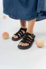 Sandalia estilo gladiadora de color negro, confeccionada en material gamuzado sintético. Presenta múltiples tiras anchas sobre el empeine, suela plana antideslizante y ajuste lateral mediante hebilla metálica.