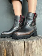 Bota de cuero con efecto desgastado en tonos marrones y negros, con suela track robusta y cierre lateral. Presenta detalles en color rojo en la parte superior de la caña.