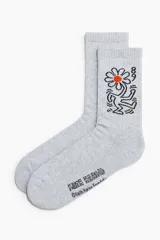 Medias de caña alta gris melange con diseño estampado estilo Keith Haring de una flor con centro naranja y figura bailando, además de la inscripción de la marca en la planta.