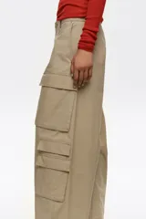 Pantalón cargo beige de tiro bajo y corte ancho, con múltiples bolsillos con solapa en los laterales de las piernas y cierre frontal con botón y cremallera oculta.