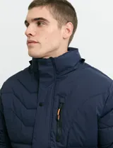 Campera acolchada de color gris, con capucha desmontable, cierre frontal y bolsillos laterales con cremallera. Presenta un diseño de costuras horizontales y detalles en color naranja en los tiradores de los cierres.