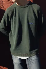 Buzo de punto acanalado color verde oscuro, de manga larga con diseño raglán, cuello redondo y un pequeño parche bordado en el pecho. Presenta detalles de rayas en los puños y el ruedo.