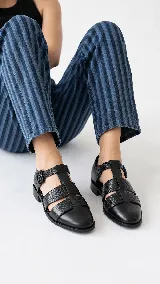 Sandalias romanas cerradas de cuero negro con textura grabada, puntera almendrada, taco bajo y tira con hebilla en el tobillo.