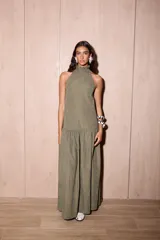 Vestido largo holgado color verde oliva, con escote halter y cuello alto con volado. Presenta un corte con fruncido a la altura de la cadera que genera un maxi volado en la falda.