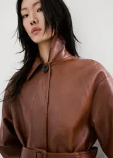Chaqueta larga de efecto cuero color marrón, con cuello camisero, botonadura frontal oculta, bolsillos laterales y cinturón ajustable con hebilla en la cintura. Cuenta con trabillas en las muñecas y diseño amplio.