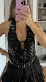 Top negro con escote halter profundo, decorado con bordados de lentejuelas y canutillos plateados en patrones geométricos y florales. Presenta un diseño con caída fluida y una pequeña tira horizontal en el centro del escote.