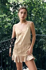 Vestido mini de tejido satinado color beige, con escote en pico adornado con encaje, tirantes anchos y detalle de encaje bordado en la cintura. El bajo presenta un acabado con volantes.