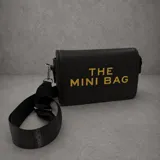 Cartera tipo bandolera de color negro con textura granulada, presenta la inscripción 'THE MINI BAG' en letras doradas en el frente. Incluye una correa ancha ajustable y extraíble.