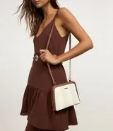 Cartera bandolera pequeña con estampado de rayas verticales en tonos beige y blanco, con detalles en marrón. Tiene cierre de cremallera, asa de cadena dorada y correa de cuero sintético marrón.