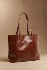 Bolso tipo tote bag color marrón, confeccionado en cuero sintético, con doble asa superior y detalle de placa metálica dorada con logo en la parte frontal.