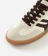 Championes de estilo urbano con diseño retro, confeccionados en material sintético color crema con detalles en marrón oscuro. Presentan recortes laterales, cierre con cordones y suela de goma color caramelo.