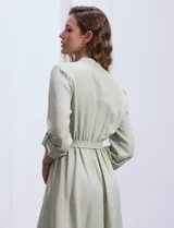 Vestido maxi color beige, con cuello mao y escote en V. Presenta mangas largas con presilla y botón para regular el largo, y un lazo ajustable en la cintura que marca la silueta.