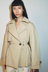 Trench corta de color beige con cuello de solapa y manga larga. Cuenta con bolsillos delanteros, cintura ajustable con cordones, bajo abullonado y cierre frontal cruzado con un botón visible. El interior está forrado con tela a cuadros.