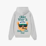 Hoodie gris de corte oversize con capucha y bolsillo frontal tipo canguro. Presenta un estampado gráfico en la espalda de un oso con anteojos de sol, gorra y un cigarro, acompañado por la frase 'Older, Wiser & Nude Project Hotter Than Ever'.