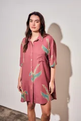 Vestido camisero corto de color rosado con estampado de aves abstractas en tonos verdes y azules. Cuenta con cuello camisero, cierre frontal con botones, mangas cortas y un bolsillo aplicado en el pecho.