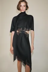 Vestido midi de satén negro con cuello subido, manga corta tipo capa y pañuelo incorporado en el mismo tejido. Presenta aberturas laterales decoradas con encaje a tono y bajo asimétrico con detalles de encaje. Cierre en la espalda con abertura y botones forrados.