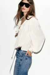 Blusa de georgette en color blanco, con escote en pico, detalle de volantes y lazada frontal. Presenta mangas largas abullonadas con puños ajustados y un bajo asimétrico con diseño tipo globo.