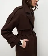 Trench coat largo color marrón chocolate, confeccionado en tejido tipo paño, con solapas amplias, cierre frontal cruzado con cinturón de la misma tela para anudar y detalles de trabillas en los hombros y puños.