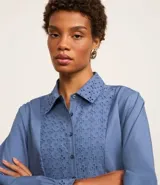 Vestido corto de corte evasé confeccionado en tricolina de algodón, color azul. Presenta cuello camisero, cierre frontal de botones y un panel central con detalle bordado calado. Posee mangas largas con puños abotonados.