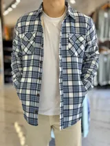Sobrecamisa de hombre a cuadros en tonos celeste, blanco y azul marino, con forro interno de corderito, bolsillos en el pecho y cierre frontal con botones.