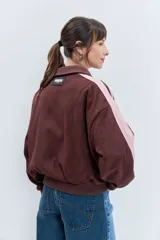 Cazadora de estilo deportivo en color marrón, confeccionada en tejido tipo gamuza. Presenta cierre frontal completo, cuello alto, puños y ruedo acanalados, y franjas laterales en tono rosado. Incluye el logo de la marca bordado en el pecho.
