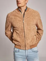 Campera bomber color marrón claro confeccionada en gamuza, con cierre frontal y bolsillos laterales con botón a presión. Presenta cuello, puños y cintura acanalados.