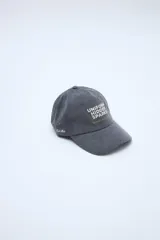 Gorra gris con visera curva, bordado frontal con texto en color beige y detalle bordado lateral.