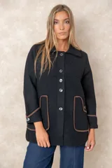 Chaqueta negra de corte amplio y diseño estructurado, con cierre frontal de botones. Presenta bolsillos frontales y puños con ribetes en contraste color marrón.