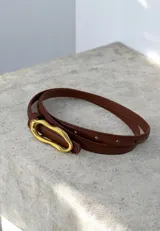 Cinto fino de cuero color marrón con hebilla metálica irregular en tono dorado.