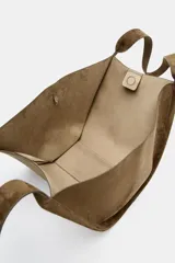 Cartera shopper de hombro confeccionada en gamuja color marrón, con diseño estructurado de laterales abiertos y doble asa superior. Cuenta con cierre de botón imantado e incluye un bolsillo interior extraíble con cremallera.