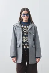Tapado gris con capucha, cierre frontal con cremallera, bolsillos laterales con solapa y bolsillo superior con cierre. Presenta un diseño estructurado de líneas modernas.
