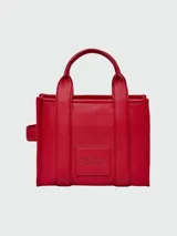 Cartera tipo tote de cuero rojo de la marca Marc Jacobs, modelo The Leather Small Tote Bag. Presenta doble asa superior, correa ajustable y extraíble para el hombro, y el texto grabado en relieve "THE TOTE BAG MARC JACOBS" en el frente.