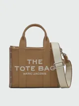 Bolso tipo tote Marc Jacobs modelo The Small Tote, color beige con detalles y texto en negro que dice "THE TOTE BAG MARC JACOBS". Cuenta con asas superiores y laterales para llevar en la mano o al hombro.