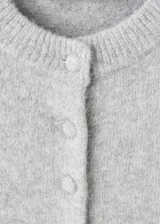 Cardigan de punto gris con cuello redondo, cierre frontal de botones y mangas largas con terminación en volados. Presenta un diseño recto con detalle de volante en el ruedo.
