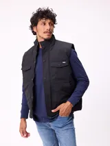 Chaleco acolchado color negro con cierre frontal de cremallera y botones a presión. Presenta cuello alto, bolsillos frontales con solapa y un pequeño parche con logo en el pecho.