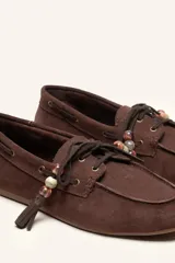 Mocasín estilo náutico de gamuza marrón, con cordones de cuero, borla decorativa y cuentas de colores en el empeine.