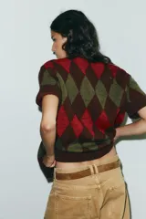 Chaqueta de punto de manga corta con cuello polo y cierre frontal de botones. Presenta un patrón de rombos (argyle) en tonos marrón oscuro, verde oliva y rojo.