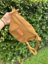 Riñonera de gamuza color tostado con cierre frontal y etiqueta de cuero con logo de la marca Antonia Wool Wear. Cuenta con correa ajustable y herrajes metálicos.