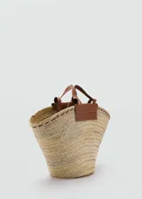 Bolso tipo cesto de tamaño grande, confeccionado en tejido de fibra natural con diseño ensanchado hacia arriba, asas largas y detalles de cuero marrón en el borde superior y la base de las asas.