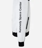 Campera tipo bomber blanca con detalles en negro, inspirada en uniformes espaciales de la NASA. Presenta parches con el logo de la NASA, la bandera de EE. UU. y texto en la manga que dice "Kennedy Space Center".
