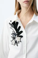 Camisa de corte recto con cuello clásico y cierre frontal de botones. Presenta un parche bordado con mostacillas en tonos blanco y negro en el frente, con diseño de estrellas y formas abstractas.