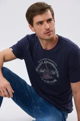 Remera azul marino de manga corta con cuello redondo, confeccionada en mezcla de algodón y poliéster. Presenta un estampado frontal circular de inspiración vintage con diseño de velero y tipografía alusiva a clubes deportivos.