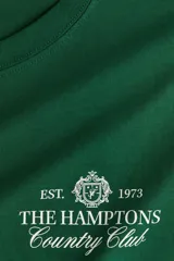 Remera de manga corta, cuello redondo y corte holgado, color verde oscuro, con estampado frontal en blanco que dice "EST. 1973 THE HAMPTONS Country Club".