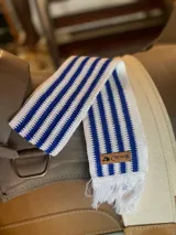 Faja de tejido horizontal a rayas blancas y azules con flecos en los extremos y aplique de cuero marrón con logo grabado.