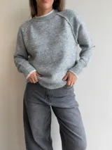 Sweater de punto de corte holgado en color gris melange, con cuello redondo y puños acanalados que presentan un detalle de contraste en color blanco. Posee mangas tipo raglán y una textura suave y elástica.